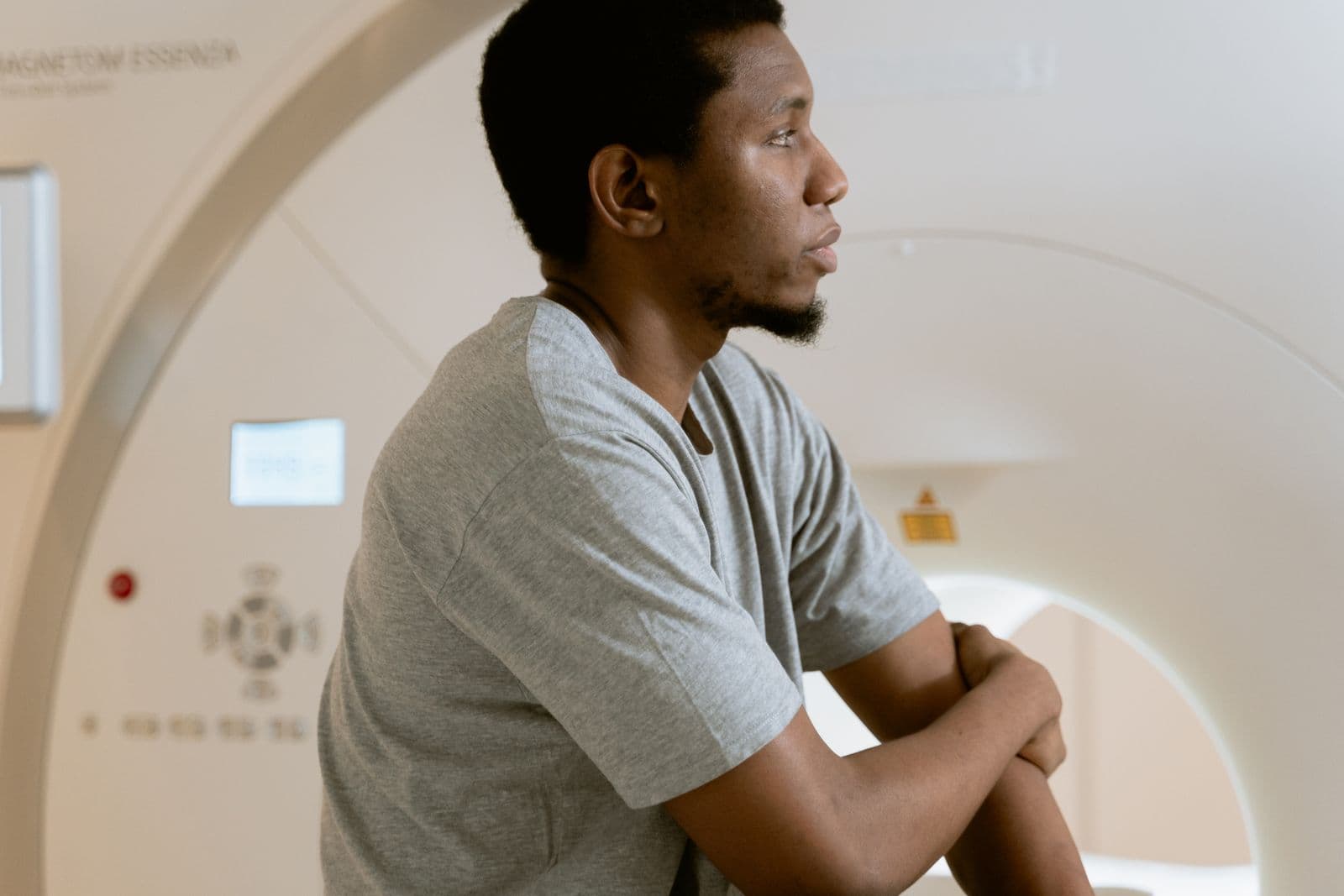 A man in a grey T-shirt waits beside an MRI scanner, looking ahead.